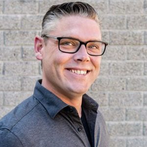 Een man met kort, netjes gestyled haar en een bril glimlacht terwijl hij voor een lichtgrijze bakstenen muur staat. Hij draagt een donker overhemd met kraag en kijkt een beetje opzij, wat de professionele uitstraling van voip den haag weerspiegelt.